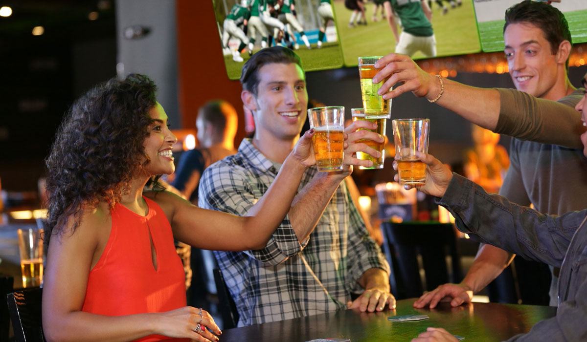 Four Customers Cheers Beer Glasses Inside A Dave & Buster's With Sports Playing On TVs In The Background