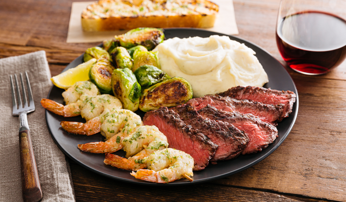 A Plate With Shrimp, Steak, Brussels Sprouts, Mashed Potatoes, And Garlic Bread Toast On The Side With A Glass Of Red Wine