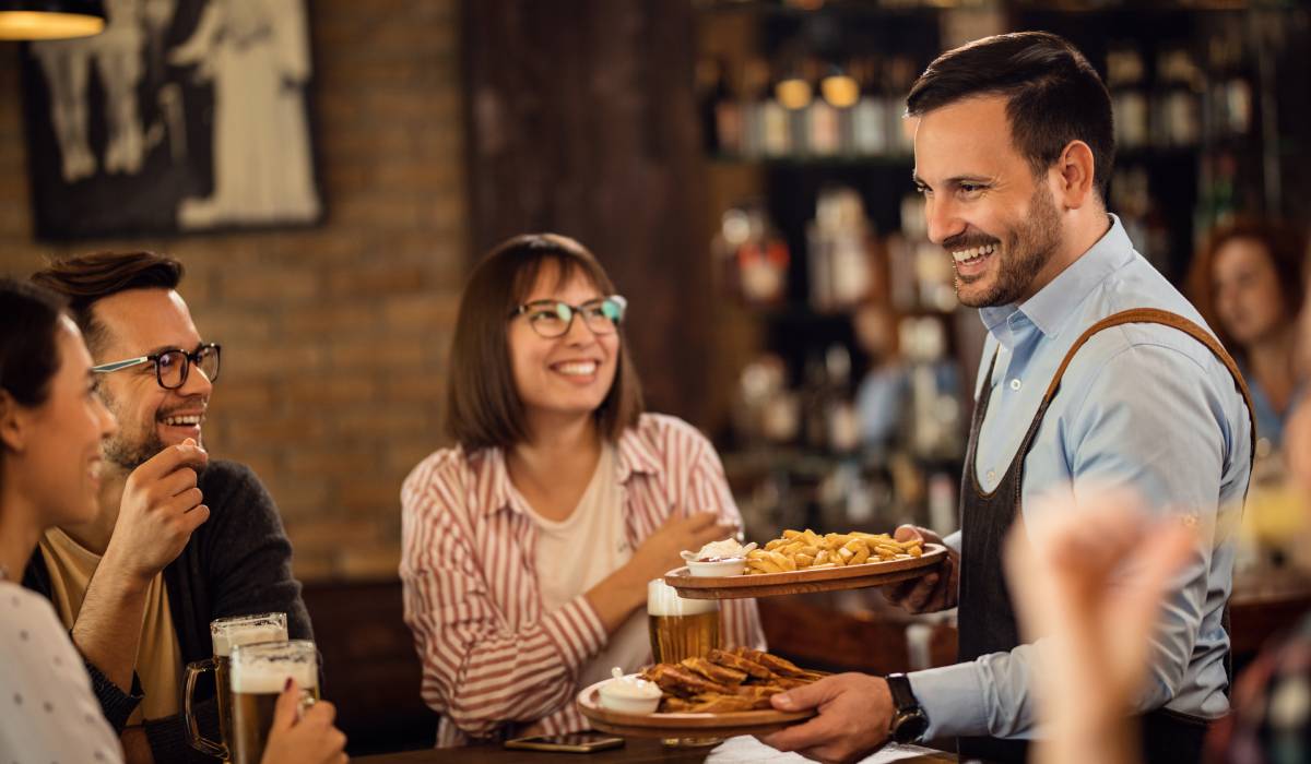 Happy Waiter Serving Food To Guest Who Are Drinking Beer In A Pub