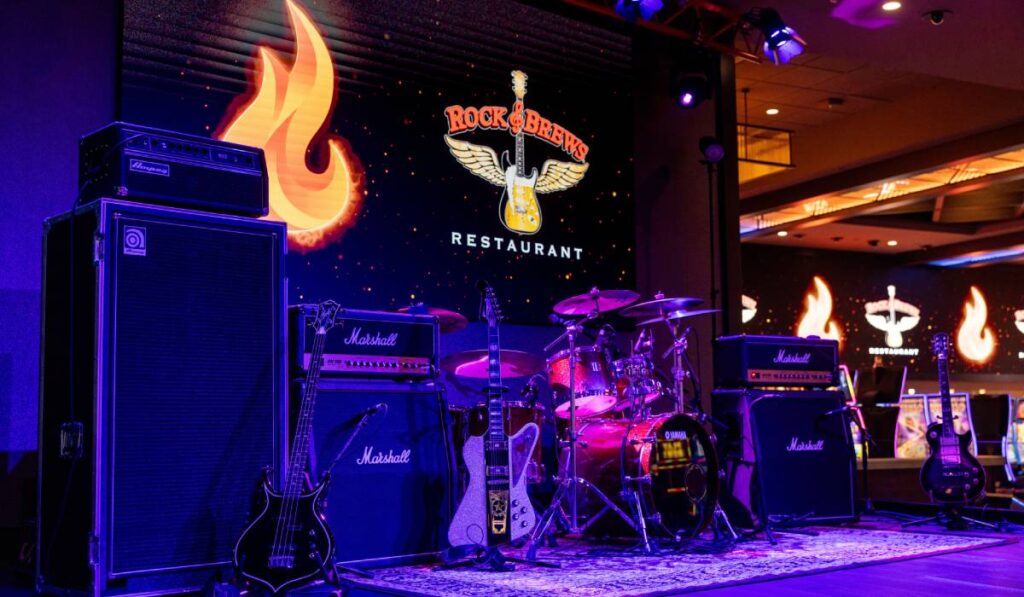 Rock & Brews stage with guitars, amps, drums, and a logo with a guitar and wings.