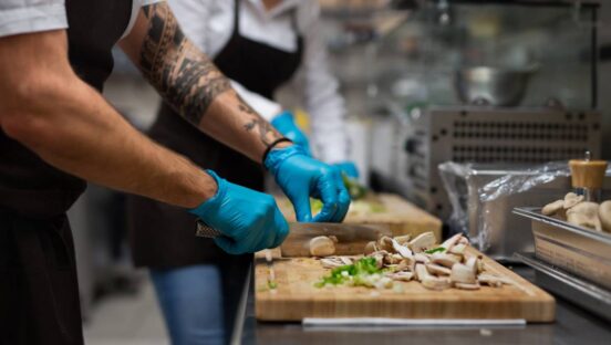 A chef with gloves cutting vegetables indoors in restaurant kitchen.